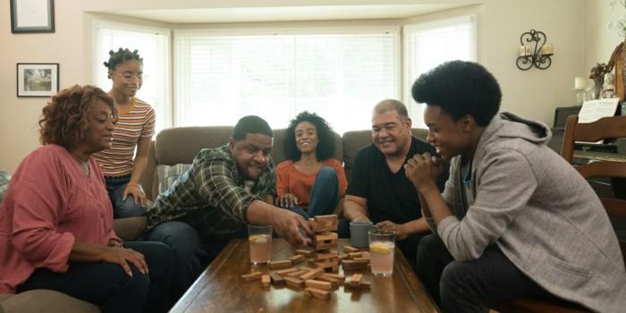 6 miembros de una familia afroamericana ríen mientras juegan Jenga en la sala de la casa.